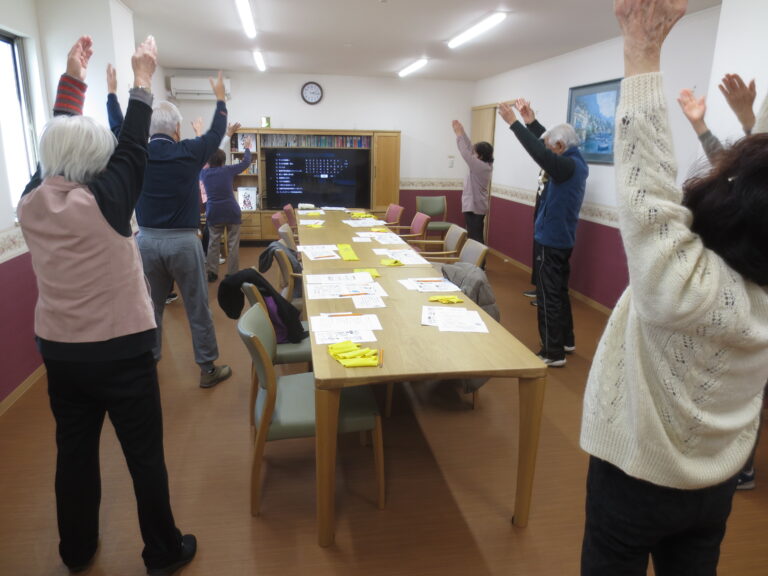 介護施設での高齢者向けレクリエーション。複数の入居者が腕を上げて体操に参加している。